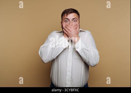 Ein Mann steht vor einem schlichten beigen Hintergrund und erscheint schockiert, als er seinen Mund mit beiden Händen bedeckt. Seine weiten Augen drücken Überraschung und Unglauben aus Stockfoto