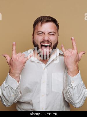 Ein Mann, der ein weißes Hemd trägt, macht einen verspielten Ausdruck, indem er seine Zunge herauszieht und das Felsenzeichen mit seinen Händen zeigt. Er hat einen Bart und ist posi Stockfoto