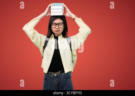 Junge Frau mit Rucksack, die einen Stapel Bücher auf dem Kopf vor rotem Hintergrund balanciert Stockfoto