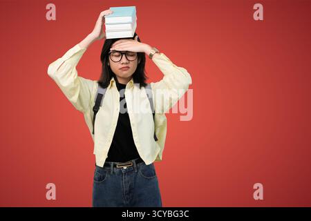Junge Frau mit Rucksack, die einen Stapel Bücher auf dem Kopf vor rotem Hintergrund balanciert Stockfoto