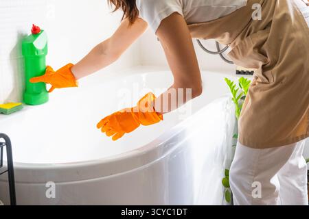 Frau in Gummihandschuhen und Schürze wäscht die Badewanne manuell im Badezimmer des Hauses mit Schwammreiniger und Reinigungsmittel in einer Flasche. Auftrag wird wiederhergestellt, Stockfoto