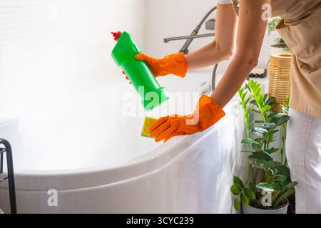 Frau in Gummihandschuhen und Schürze wäscht die Badewanne manuell im Badezimmer des Hauses mit Schwammreiniger und Reinigungsmittel in einer Flasche. Auftrag wird wiederhergestellt, Stockfoto