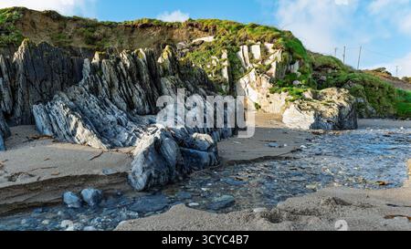 Klares Wasser fließt über Felsen an einem Sandstrand in Sheep Cove, Irland. Interessante Felsformationen treffen auf eine grasbewachsene Klippe unter einem blauen, teilweise bewölkten Himmel. Stockfoto