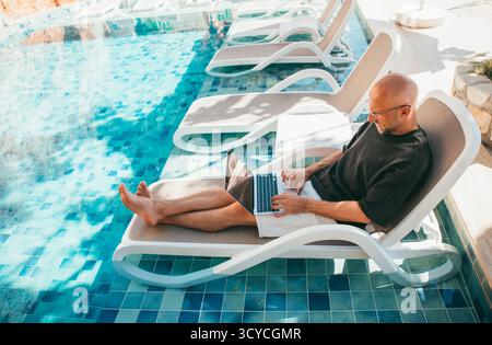Freiberuflicher Geschäftsinhaber mittleren Alters, der seinen Laptop auf einer Sonnenliege am Pool des Luxushotels arbeitet. Digital Nomad leitet erfolgreich das Unternehmen Re Stockfoto
