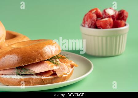 Ein Bagel mit Lachs und Dill ist auf einem weißen Teller. Neben dem Sandwich gibt es eine Schüssel mit Kirschtomaten Stockfoto
