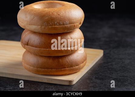 Drei Bagels, die auf einem Holzschneidebrett übereinander gestapelt sind. Die Bagels sind braun und scheinen frisch gebacken zu sein. Konzept von Genuss und Co Stockfoto