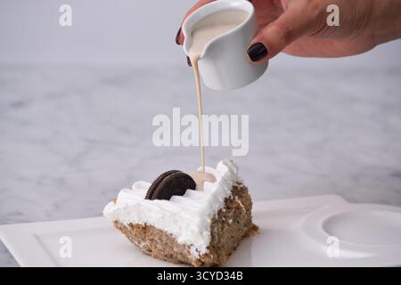 Oreo Cream Cake Slice mit zerkleinerten Schokoladen-Sandwichkeksen, garniert mit Schlagsahne und tropfender Milchsauce, serviert auf einem Teller, der auf Background isoliert wird Stockfoto