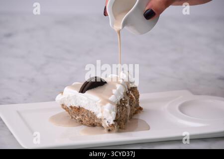 Oreo Cream Cake Slice mit zerkleinerten Schokoladen-Sandwichkeksen, garniert mit Schlagsahne und tropfender Milchsauce, serviert auf einem Teller, der auf Background isoliert wird Stockfoto