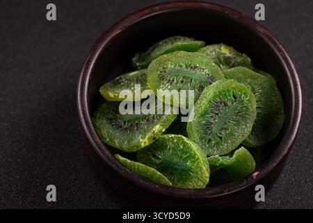 Getrocknete Kiwi-Fruchtscheiben mit leuchtend grünen Farben und sichtbaren schwarzen Samen füllen eine dunkle Keramikschale auf einem strukturierten Hintergrund, die ein darstellt Stockfoto