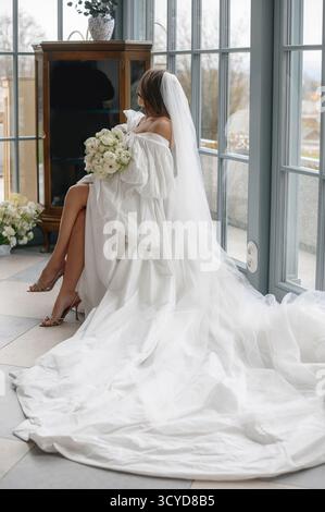 Braut in elegantem weißem Kleid mit langem Zug, Blumenstrauß in der Hand, sitzt an großen Fenstern in einem hellen Raum. Stockfoto