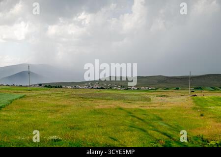 Weite Steppenlandschaft mit grünen Feldern, fernen Bergen und einem kleinen Dorf, das im Hintergrund zu sehen ist. Javakheti, Georgien. 18. Juli 2020. Stockfoto