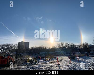 Sonnenschein und Sonnenhunde über einer schneebedeckten Baustelle mit Maschinen und urbanem Hintergrund. Stockfoto