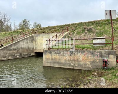 Betonabflusskanal mit rotem Geländer und Wasserfluss darunter, Teil der Regenwasser- oder Bewässerungsinfrastruktur in einem ländlichen Gebiet. Stockfoto