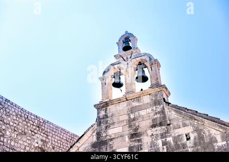 Steinglockenturm einer historischen Kirche in der Altstadt von Dubrovnik, Kroatien, mit zwei Bronzeglocken, die vor einem hellen Sommerhimmel eingerahmt sind. Stockfoto