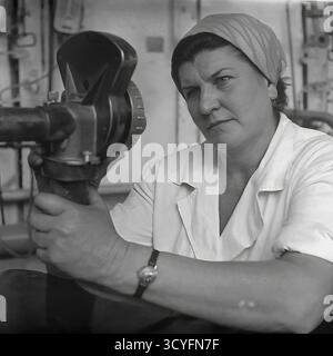 Dieses Archivfoto aus Sloviansk, UdSSR, aus den 1970er Jahren, ist ein Porträt einer Arbeiterin der Slawjansk Armature-Isolator Plant (AIZ). Mit einem weißen Mantel und einem Kopftuch (Platok) hält sie selbstbewusst ein komplexes technisches Gerät und blickt direkt in die Kamera. Dieses quadratische Bild zeigt das qualifizierte und engagierte Gesicht der sowjetischen Industriearbeiter, möglicherweise in einem Qualitätskontrolllabor Stockfoto