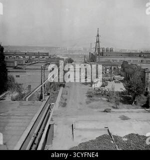 Dieses archivierte Schwarzweißfoto aus den 1970er Jahren bietet eine vertikale Panoramaansicht der Slawjansk Armature-Isolator Plant (AIZ) in Sloviansk, UdSSR. Der Schuss blickt auf einen großen Fabrikhof, flankiert von Backsteinwerkstätten. Ein markantes Merkmal ist ein großer elektrischer Übertragungsmast (Power Tower), der auf dem Werksgelände neben erhöhten Rohrleitungen steht. Im Vordergrund stapeln sich fertige Isolatoren. In der Ferne sind die Wohngebäude der Stadt sichtbar. Stockfoto