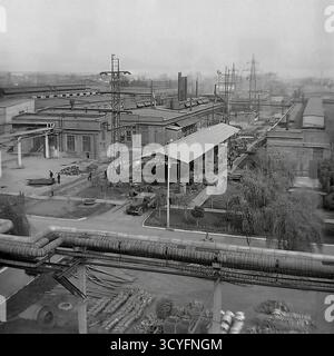 Dieses archivierte Schwarzweißfoto aus den 1970er Jahren bietet einen Panoramablick auf die Slawjansk Armature-Isolator Plant (AIZ) in Sloviansk, UdSSR. Die Aufnahme zeigt die riesige Industrielandschaft. Im Vordergrund befindet sich eine große, erhöhte Pipeline. Unterhalb des Werks befinden sich ein Lkw (Typ GAZ-69), ein langer Schuppen und Arbeiter. Der Hintergrund ist gefüllt mit Werkstätten, hohen Schornsteinen und vielen elektrischen Übertragungsmasten (Strommasten). Stockfoto