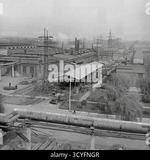 Dieses archivierte Schwarzweißfoto aus den 1970er Jahren bietet einen Panoramablick auf die Slawjansk Armature-Isolator Plant (AIZ) in Sloviansk, UdSSR. Die Aufnahme zeigt die riesige Industrielandschaft. Im Vordergrund befindet sich eine große, erhöhte Pipeline. Unterhalb des Werks befinden sich ein Lkw (Typ GAZ-69), ein langer Schuppen und Arbeiter. Der Hintergrund ist gefüllt mit Werkstätten, hohen Schornsteinen und vielen elektrischen Übertragungsmasten (Strommasten). Stockfoto