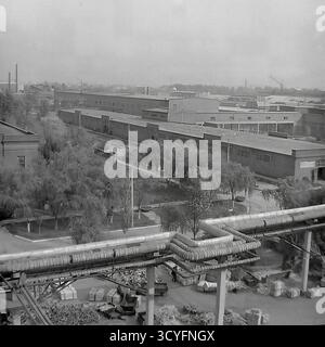 Dieses archivierte Schwarzweißfoto aus den 1970er Jahren bietet einen Panoramablick auf die Slawjansk Armature-Isolator Plant (AIZ) in Sloviansk, UdSSR. Die Aufnahme fängt die riesige Industrielandschaft ein, die im Vordergrund von einem großen Netz hochgezogener, isolierter Pipelines dominiert wird. Unterhalb des Fabrikhofs befinden sich landschaftlich angelegte Gassen mit vielen Weidenbäumen, Backsteinwerkstätten und Lagerbereiche mit Holzkisten. Im Hintergrund stehen weitere Werkstätten und Schornsteine. Stockfoto