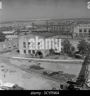 Dieses archivierte Schwarzweißfoto aus den 1970er Jahren bietet eine vertikale Hochwinkelansicht der Slavjansk Armature-Isolator Plant (AIZ) in Sloviansk, UdSSR. Der Schuss erfasst den riesigen Industriehof, der als Freiluftlager genutzt wird. Tausende fertiger Isolatoren stapeln sich auf dem Boden neben den Bahngleisen. In der Mitte befindet sich eine große Ziegelwerkstatt. Im Hintergrund erstreckt sich der Fabrikkomplex bis zur Skyline der Stadt, mit einem Gebäude, das den Slogan „Entscheidungen des 23. Kongresses der KPdSU – ins Leben!“ trägt. Stockfoto