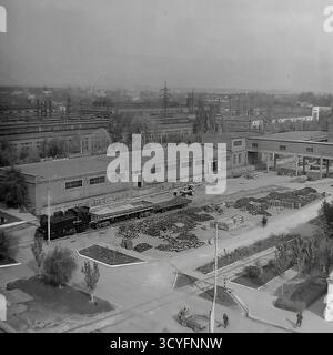 Dieses archivierte Schwarzweißfoto aus den 1970er Jahren zeigt einen Hochwinkel-Panoramablick auf die Slavjansk Armature-Isolator Plant (AIZ) in Sloviansk, UdSSR. Der riesige Industriehof dient als Freiluftlager, gefüllt mit massiven Häufern fertiger Isolatoren. Die Bilder dokumentieren die Logistik der Fabrik und zeigen eine kleine Rangierlokomotive (Nr. 8223) auf dem Gleisanschluss sowie schwere Gabelstapler, die Güter transportieren. Das ist ein Dokument sowjetischen industriellen Ausmaßes Stockfoto