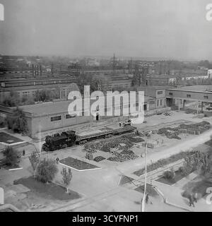 Dieses archivierte Schwarzweißfoto aus den 1970er Jahren zeigt einen Hochwinkel-Panoramablick auf die Slavjansk Armature-Isolator Plant (AIZ) in Sloviansk, UdSSR. Der riesige Industriehof dient als Freiluftlager, gefüllt mit massiven Häufern fertiger Isolatoren. Die Bilder dokumentieren die Logistik der Fabrik und zeigen eine kleine Rangierlokomotive (Nr. 8223) auf dem Gleisanschluss sowie schwere Gabelstapler, die Güter transportieren. Das ist ein Dokument sowjetischen industriellen Ausmaßes Stockfoto
