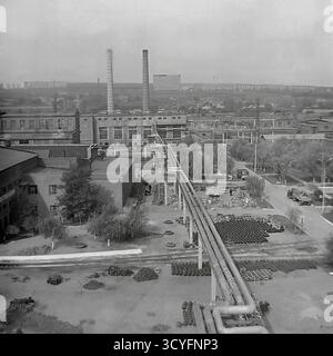 Dieses archivierte Schwarzweißfoto aus den 1970er Jahren bietet einen Panoramablick auf die Slawjansk Armature-Isolator Plant (AIZ) in Sloviansk, UdSSR. Der riesige Industriehof dient als riesiges Freiluftlager, das mit Tausenden von fertigen Isolatoren auf Paletten gefüllt ist. Durch den Hof verläuft ein Netz von Hochleitungen und Eisenbahngleisen. Ein mobiler Kran und ein hoher Schornstein sind in dieser Szene sowjetischer Industrieskala zu sehen. Stockfoto
