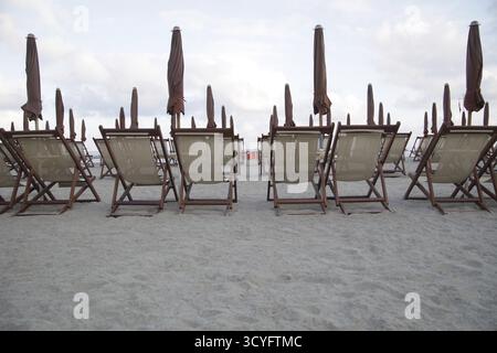 Eine Reihe von Liegestühlen steht auf dem Sand. Die Stühle sind leer und die Sonnenschirme sind geschlossen. Die Szene ist friedlich und entspannend, mit den Stühlen Stockfoto