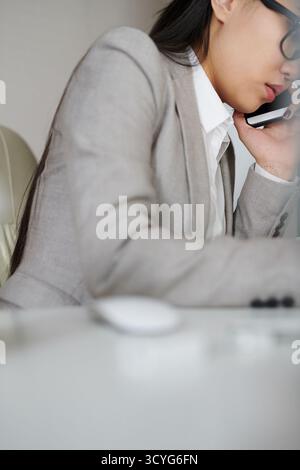 Junge asiatische Geschäftsfrau in elegantem Anzug und Brille, die auf dem Smartphone spricht, während sie am Bürotisch sitzt, mit flachem Blickwinkel Stockfoto