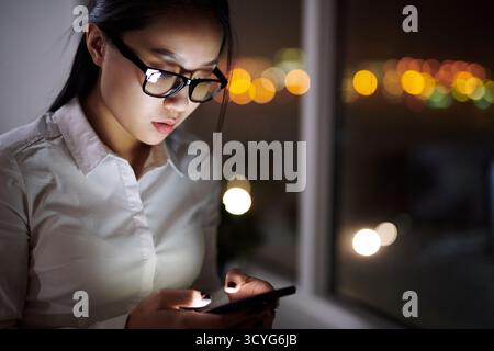 Junge asiatische Geschäftsarbeiterin, die am späten Abend am Bürofenster am Smartphone SMS schreibt, verschwommenes Licht im Hintergrund Stockfoto
