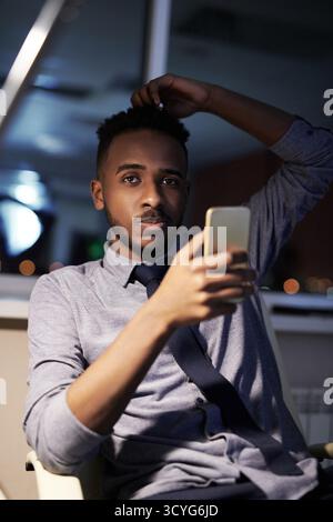 Porträt eines jungen afroamerikanischen Geschäftsmannes, der am späten Abend im Büro sitzt, Smartphone hält und in die Kamera schaut Stockfoto
