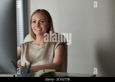 Porträt einer schönen blonden, kaukasischen Frau, die im komfortablen Büro am Tisch sitzt und fröhlich in die Kamera lächelt Stockfoto