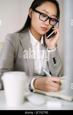 Junge asiatische Sekretärin in stilvoller Firmenkleidung, die im Büro arbeitet, am Tisch sitzt und Anrufe entgegennimmt, tiefer Blickwinkel Stockfoto