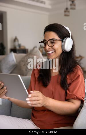 Glückliche junge hispanische Frau in Kopfhörern, die Internetkommunikation genießt Stockfoto