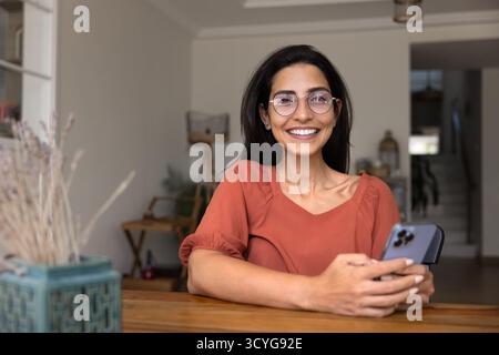 Glückliche Smartphone-Nutzerin, die zu Hause am Arbeitstisch sitzt Stockfoto