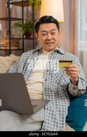 Glücklicher Mann mittleren Alters mit Kreditkarte und Laptop beim Überweisen von Geld, kauft Online-Shopping bargeldlose Bestellung Lebensmittel Lieferung zu Hause Wohnung drinnen. Der Chinese sitzt auf der Couch. Stockfoto