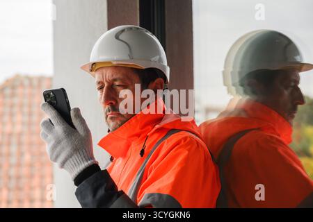 Bauingenieur mit Sicherheitsjacke und Schutzhelm mit Smartphone auf der Baustelle, Konzept der Kommunikation, Planung und moderne Technik in e Stockfoto