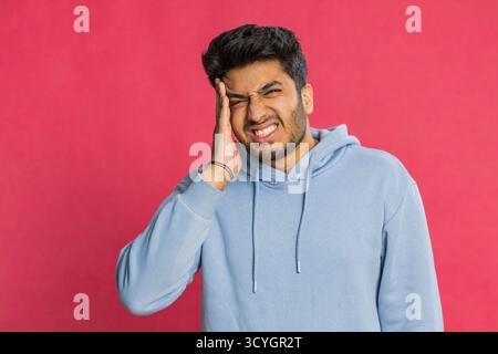 Unzufrieden erschöpfter indischer Hindu-Mann, der Schläfen reibt, um Kopfschmerzen zu heilen, leidet unter Spannung, Migräne, Stress, grimmigen Schmerzen, Bluthochdruck isoliert auf rotem Hintergrund Stockfoto