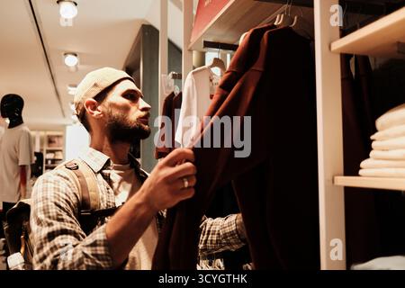 Ein junger Mann untersucht einen roten Pullover in einem modernen Bekleidungsgeschäft. Konzept von Shopping, Mode und persönlichem Stil. Stockfoto