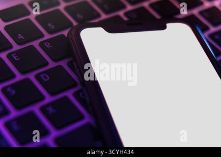 Mock Up, Smartphone mit weißem, leerem Bildschirm, das in violettem Licht auf der Laptop-Tastatur liegt, trendiges digitales Konzept für moderne Gadget-, Technologie- oder Anwendungsdesign-Projekte. Leerzeichen für Text Stockfoto