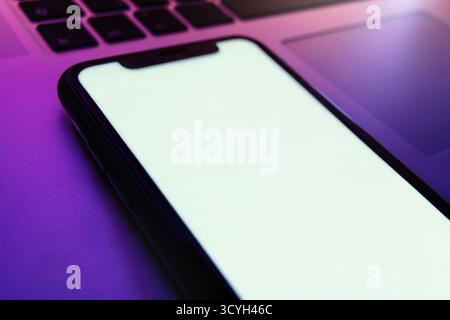Modernes Smartphone mit leerem weißen Bildschirm liegt unter lila Neonlicht auf der Laptop-Tastatur, für App-Präsentationen oder für den digitalen Hintergrund. Leerzeichen für Text Stockfoto