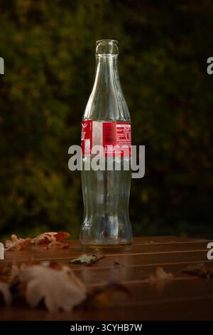 Leere Coca-Cola-Glasflasche auf einem Holztisch mit Herbstlaub in Straubing, Bayern, Deutschland. Nur Redaktionelle Verwendung. Stockfoto