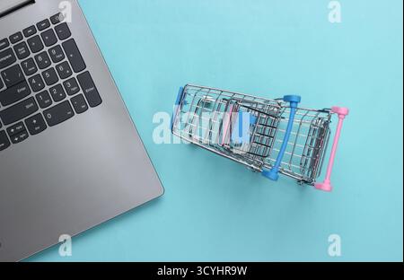 Online-Shopping. Laptop mit Supermarktwagen auf blauem Pastellhintergrund. Draufsicht. Flache Lagen Stockfoto