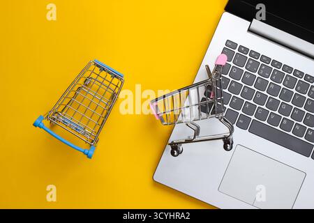 Online-Shopping. Laptop mit Supermarktwagen auf gelbem Hintergrund. Draufsicht. Flache Lagen Stockfoto