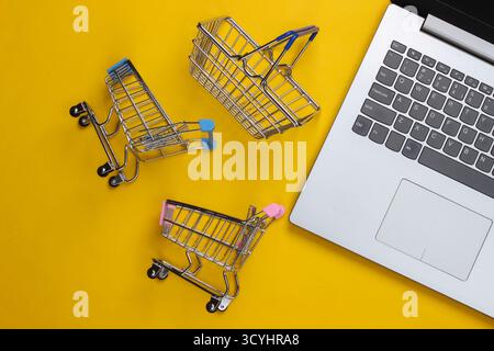 Online-Shopping. Laptop mit Supermarktwagen, Korb auf gelbem Hintergrund. Draufsicht. Flache Lagen Stockfoto
