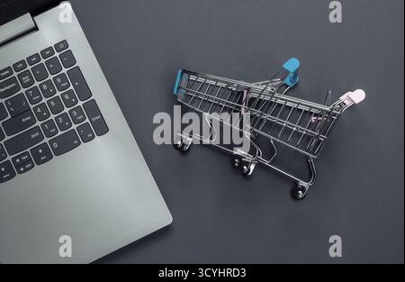 Online-Shopping. Laptop mit Supermarktwagen auf grauem Hintergrund. Draufsicht. Flache Lagen Stockfoto