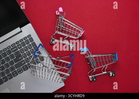 Online-Shopping. Laptop mit Supermarktwagen, Korb auf rotem Hintergrund. Draufsicht. Flache Lagen Stockfoto
