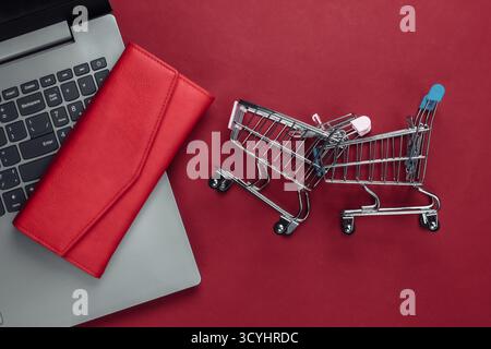 Online-Shopping. Laptop mit Supermarktwagen, Brieftasche auf rotem Hintergrund. Draufsicht. Flache Lagen Stockfoto