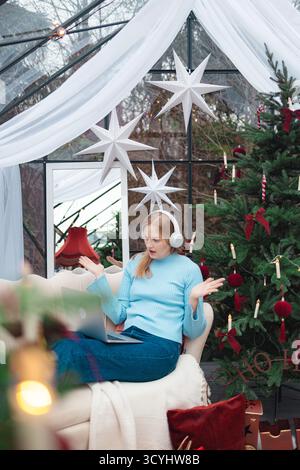 Blonde, kaukasische Frau in einem hellblauen Pullover, die auf dem Sofa neben dem geschmückten Weihnachtsbaum sitzt, ein Smartphone hält und Überraschung oder Verwirrung ausdrückt. Stockfoto