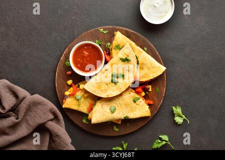 Köstliche Quesadilla mit frischem Gemüse serviert mit Sauce auf Holzplatte auf braunem Steinhintergrund. Draufsicht, flach Stockfoto